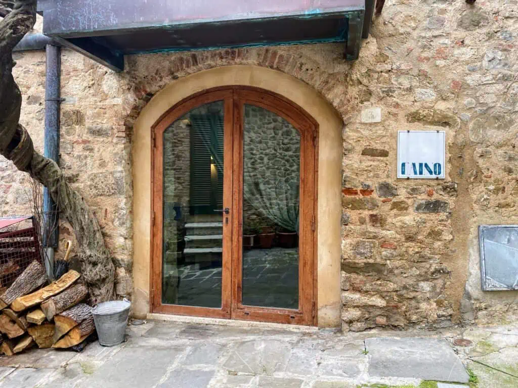 Front entrance to Caino restaurant. Large glass doors framed in wood. Wood pile on left. Stone floor outside entrance.