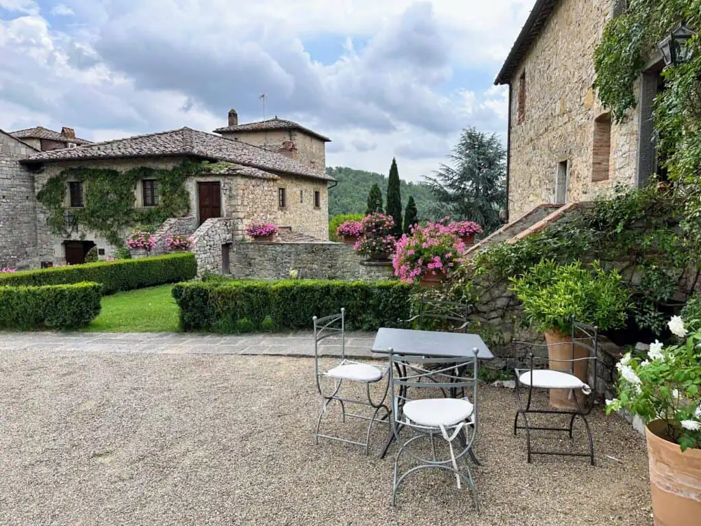 Buildings at Castello di Spaltenna hotel in Gaiole in Chianti. Stone buildings, partially covered in ivy. Gravel area in foreground with table and chairs.