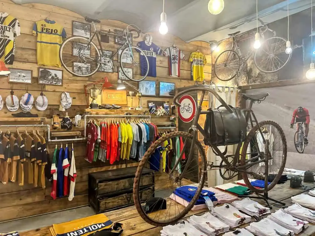 Vintage bicycle sits on a table in a shop. You can see vintage bicycle jerseys on walls and two more vintage bikes hanging from ceiling in back of photo. Jerseys and t-shirts for sale on table and on racks.