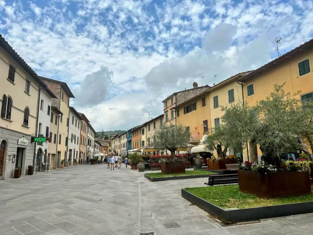 Main square in Gaiole in Chianti in Tuscany. You can see olive trees in planters on the right, people walking at the far end of the square, and colorful buildings on either side. Sunny day.