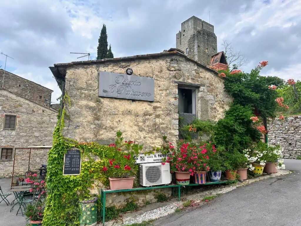 Stone building in Italy. It's the restaurant, Osteria Il Papavero, as shown on metal sign on side of wall. Plants and ivy decorate the stone walls. Tables and chairs on left side of building.