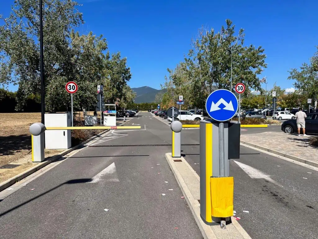 Blue sign with arrows showing drivers to enter one of the two lanes blocked by a bar. In the background you can see a large rental car parking lot that's kind of shaded by trees.