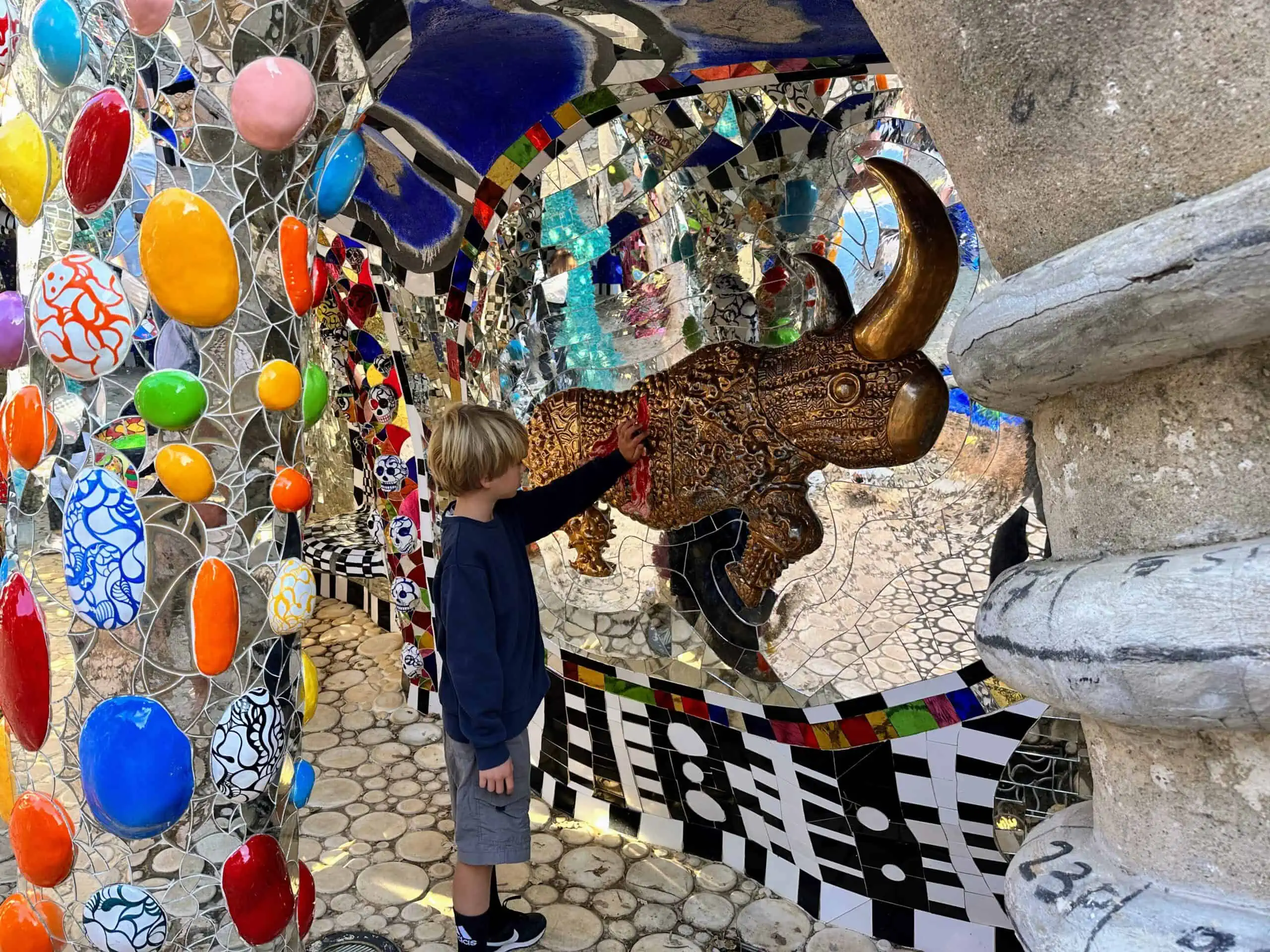 Boy touches colorful rhino sculpture in Italy. The walls and columns around are made of colorful ceramics and glass mosaics.