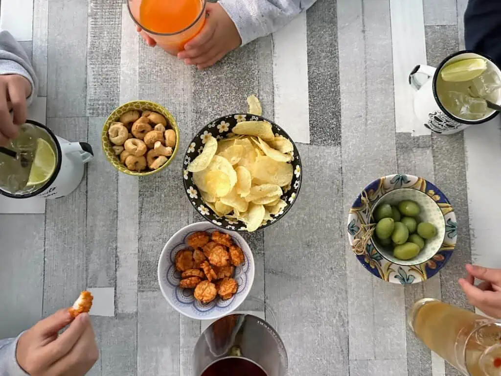 Kids hands reaching for small bowls of chips, olives, taralli and rice cakes. Also drinks on table.