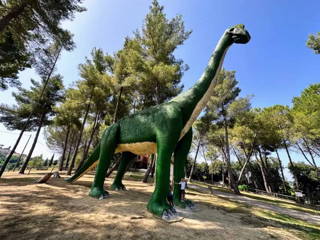 Two boys hug feet of large green dinosaur in a dirt field. Trees surround the field.
