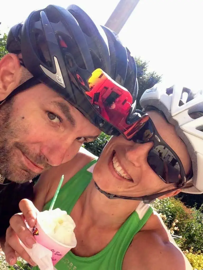 Close up of couple wearing bike helmets and looking at camera. Woman is holding cup of gelato.