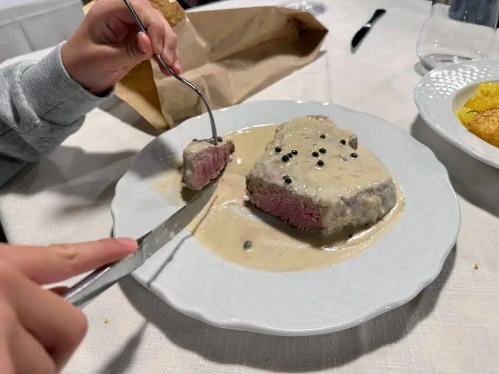 Child's hand's cutting a beef filet on a white plate. Green pepper sauce on top of the steak.