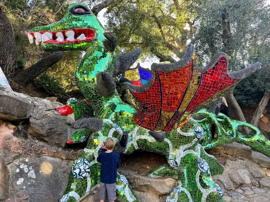 Boy looks at large green and red mosaic dragon sculpture. It's sitting in rocky, forested area.