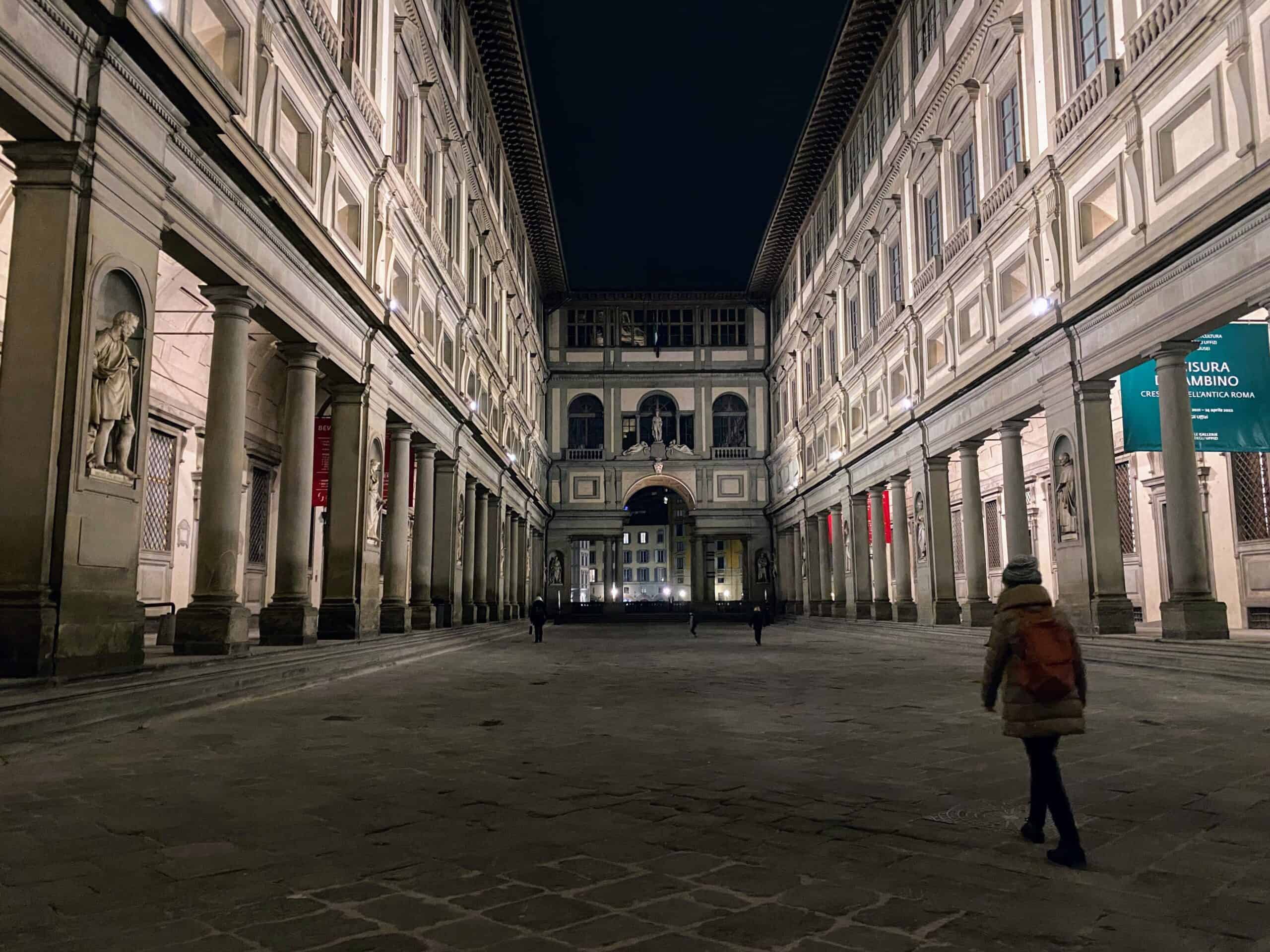 Woman in coat and hat walks in empty piazza at the Uffizi Gallery in Florence, Italy. There are three more people at the far end of the piazza. It's night.
