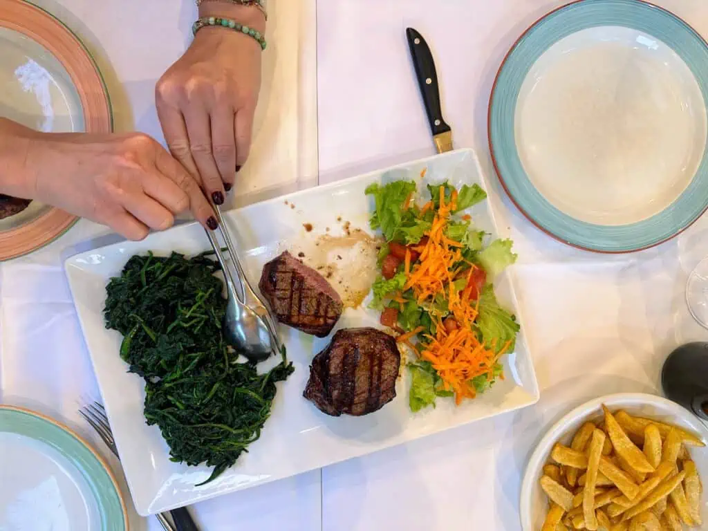 Bird's eye view of table in Florence, Italy restaurant. You can see hands serving from a plate of spinach, beef filet, and salad. Bowl of french fries in lower right.