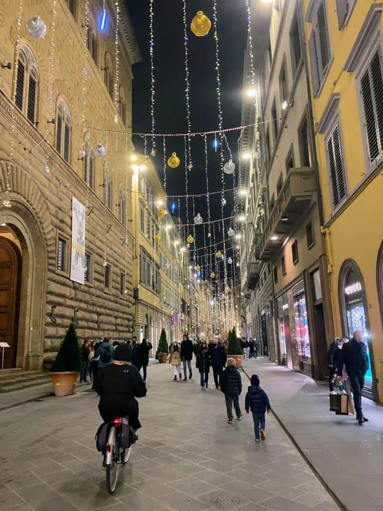 Christmas lights hang between buildings on Via Tornabuoni in Florence, Italy. People walk down the street in coats. Woman cycling in foreground.