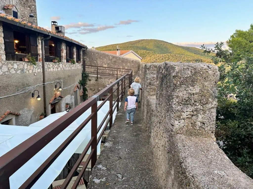 Two children walking on medieval wall of town of Capalbio in Tuscany, Italy. You can see buildings on left and hill in background.