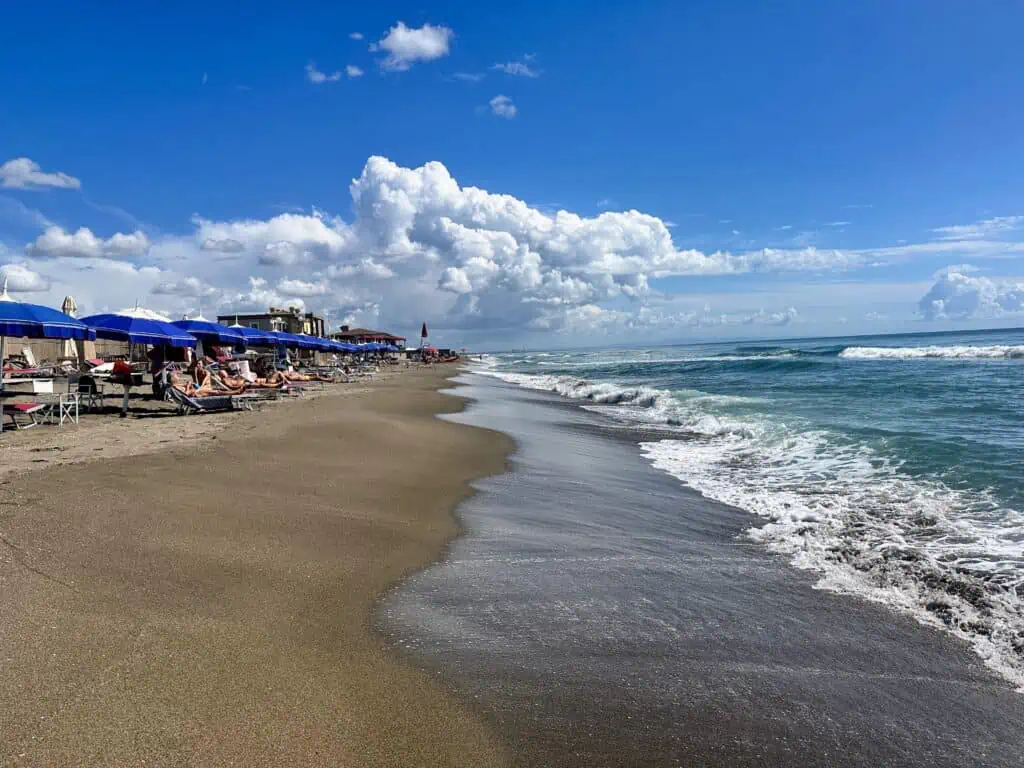 Waves lap the shore at Macchiatonda beach in Tuscany, Italy. Blue beach umbrellas and sunbathers on the left.