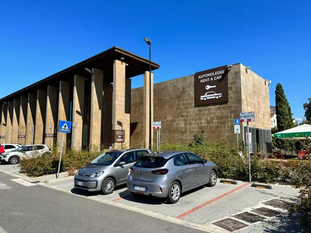Large brown brick building with brown sign for 'Autonoleggi Rent a Car' and a key and car icon. Cars parked in front of the building.