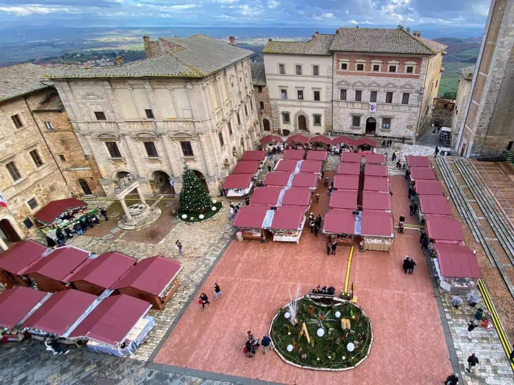 Red roofs of market stalls at the Montepulciano Christmas Market. It's in a main piazza. You can also see a large Christmas tree on left by well. Large buildings surround the piazza.