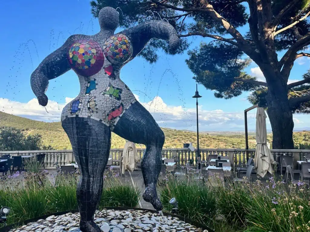 Nanà Fontaine sculpture by Niki St. Phalle. It's a colorful sculpture of a voluptuous woman made using mosaic pieces. Terrace with countryside view in background.