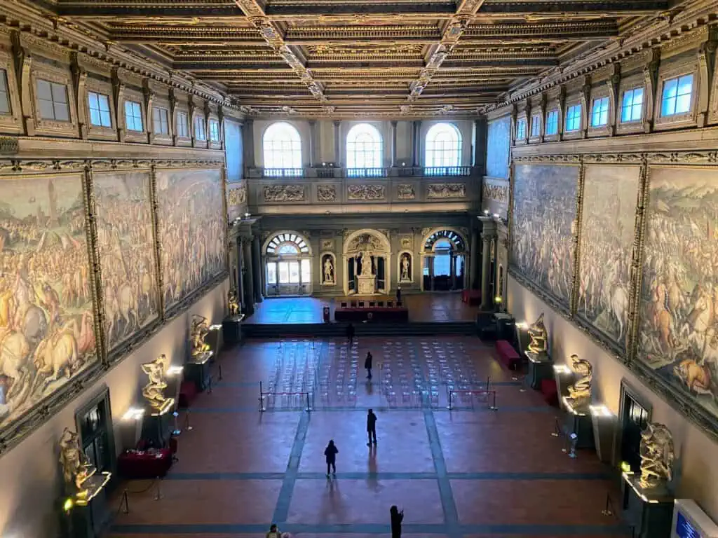 A few people are inside the Palazzo Vecchio's Hall of 500 in Florence, Italy. Massive paintings hang on the walls. Chairs are set up in the far side of the room.