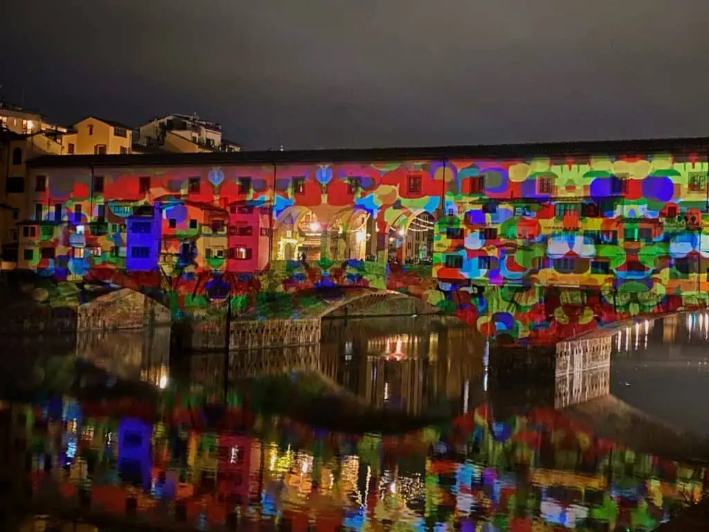 Ponte Vecchio bridge in Florence, Italy being lit up with a colorful light display at night. It's reflecting on the Arno River.