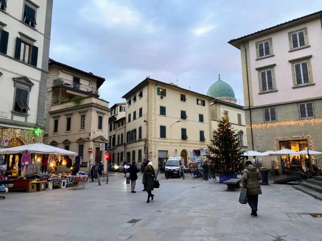 A few people walk in a square in Sant'Ambrogio in Florence, Italy. It's daytime and you can see a Christmas tree in the square and Christmas lights on a couple of the buildings.