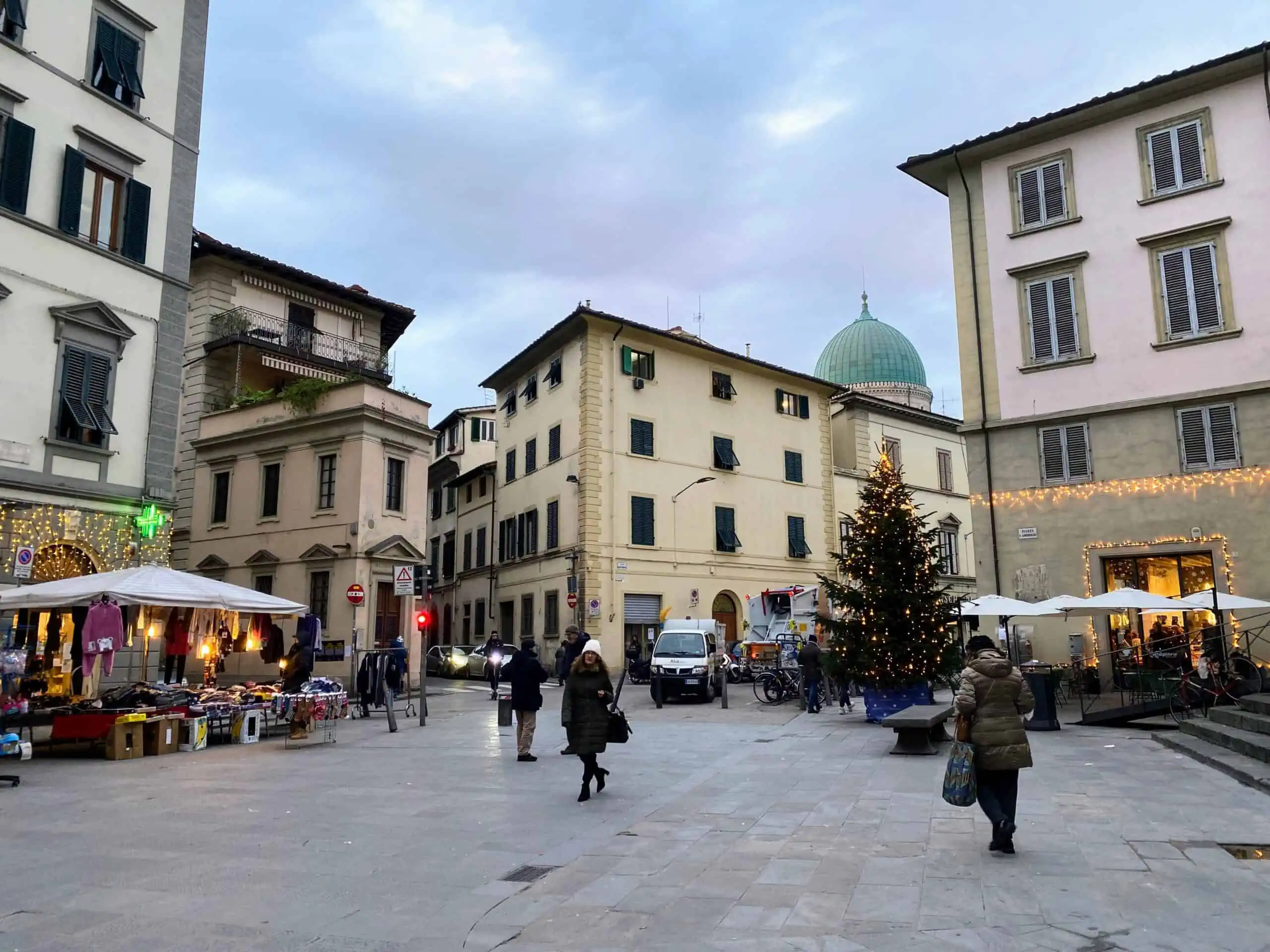A few people walk in a square in Sant'Ambrogio in Florence, Italy. It's daytime and you can see a Christmas tree in the square and Christmas lights on a couple of the buildings.