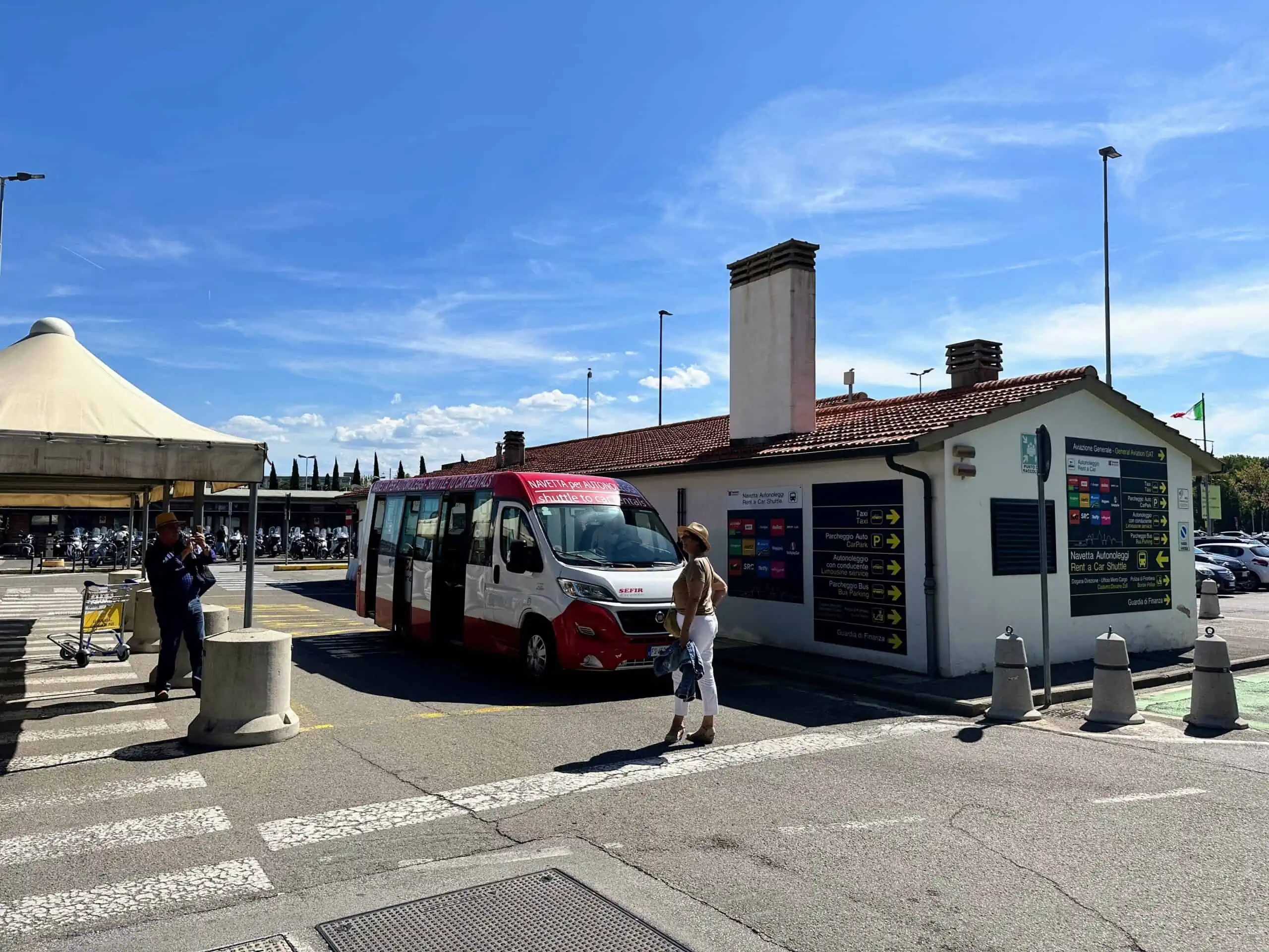 A couple is waiting next to a white and red shuttle bus at the Florence airport. There is a waiting area with a tent on the left and a small building next to the bus on the right.