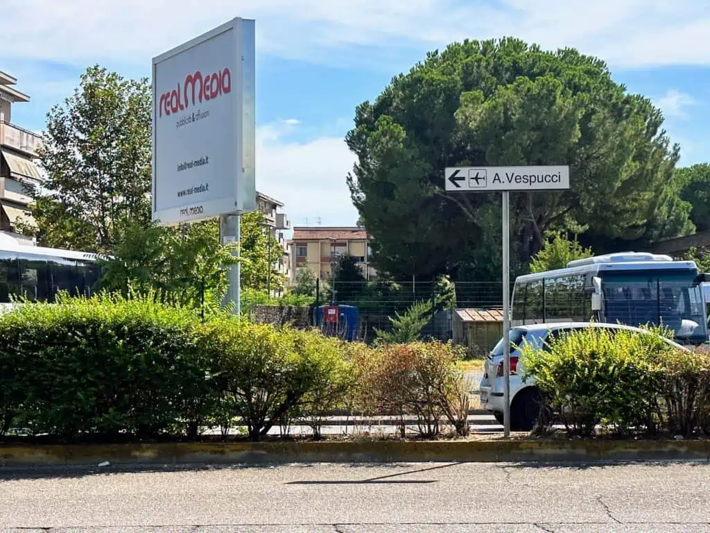 Intersection with small white sign for 'A.Vespucci' and an airplane icon. You can see a car driving on road in the background and trees and a billboard with Italian text.