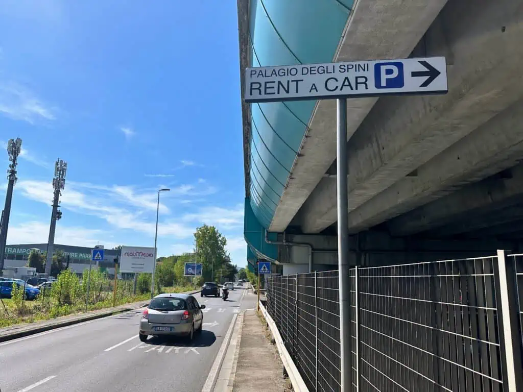 White sign pointing right toward 'Palagio degli Spini Rent a Car' with blue 'P' icon. Cars driving on road on left. Sunny day.
