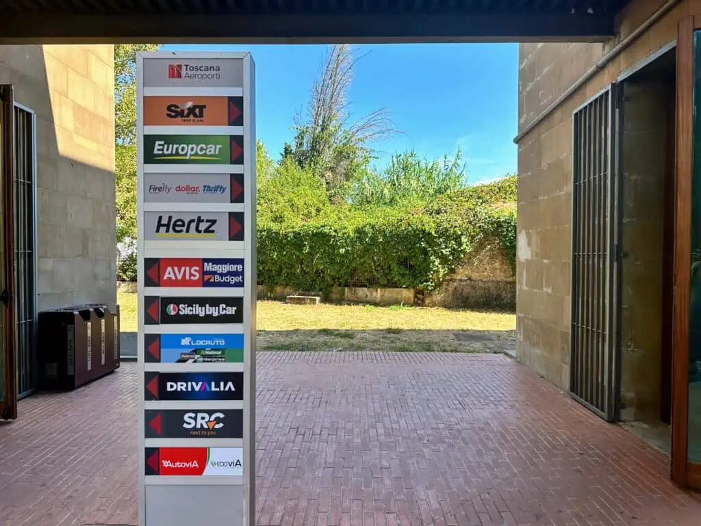 Colorful signs for car rental companies on a column in a courtyard. They have arrows that point right or left to their respective offices at the Florence airport car rental location.