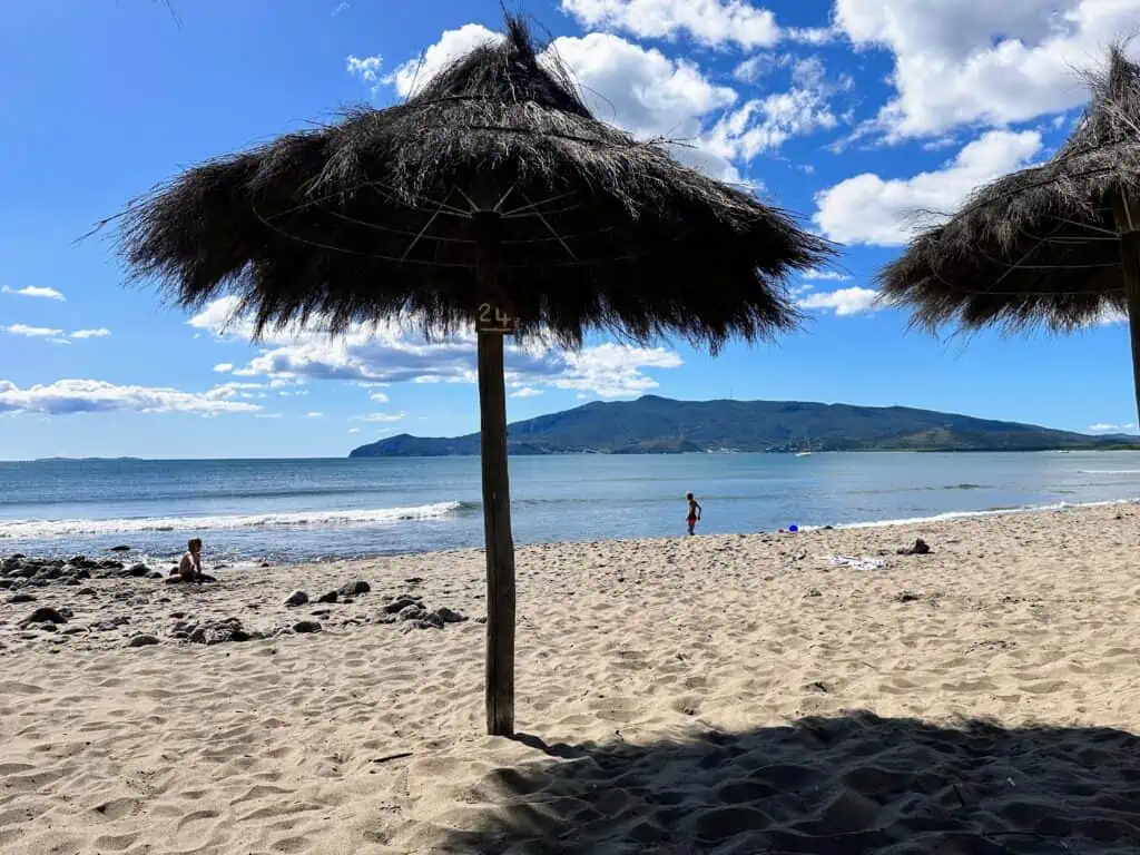 View of grassy umbrellas and kids playing in the sand at Feniglia beach in Tuscany. In the background you can see Monte Argentario.