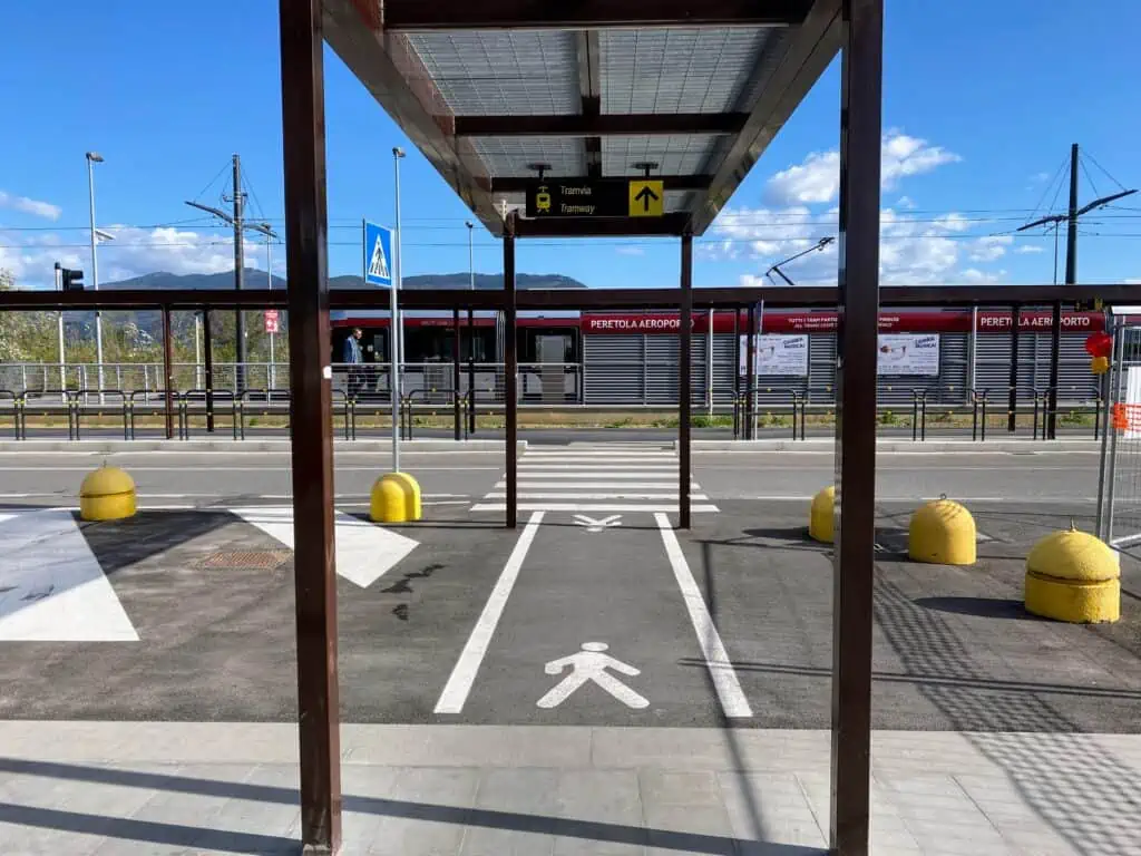 View of tram stop at the Florence airport. You can see the walkway and an overhead covering that leads to the tram stop straight ahead. There is a tram waiting and a man standing next to it. Sunny day with puffy white clouds.
