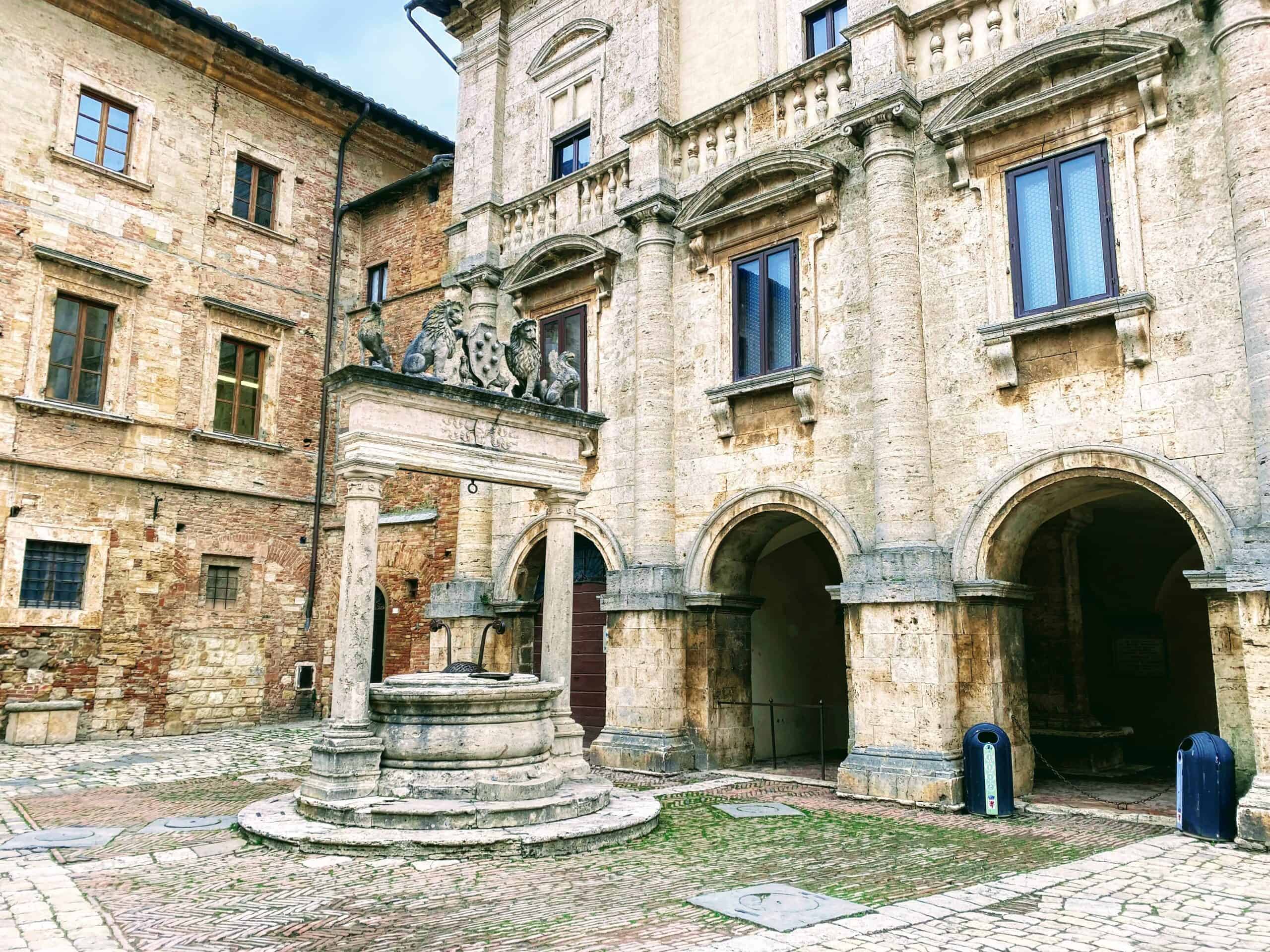 Stone well in corner of piazza. Grass growing in cracks between bricks on the ground. Lions and Medici crest with 6 balls decorate top part of well.Stone buildings in background.