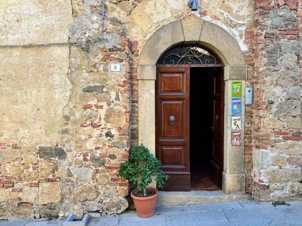 Wooden door in stone entrance to B&B La Casa di Adelina. Guidebook signs on right side of door and small plant to left of door. Old stone and brick walls.