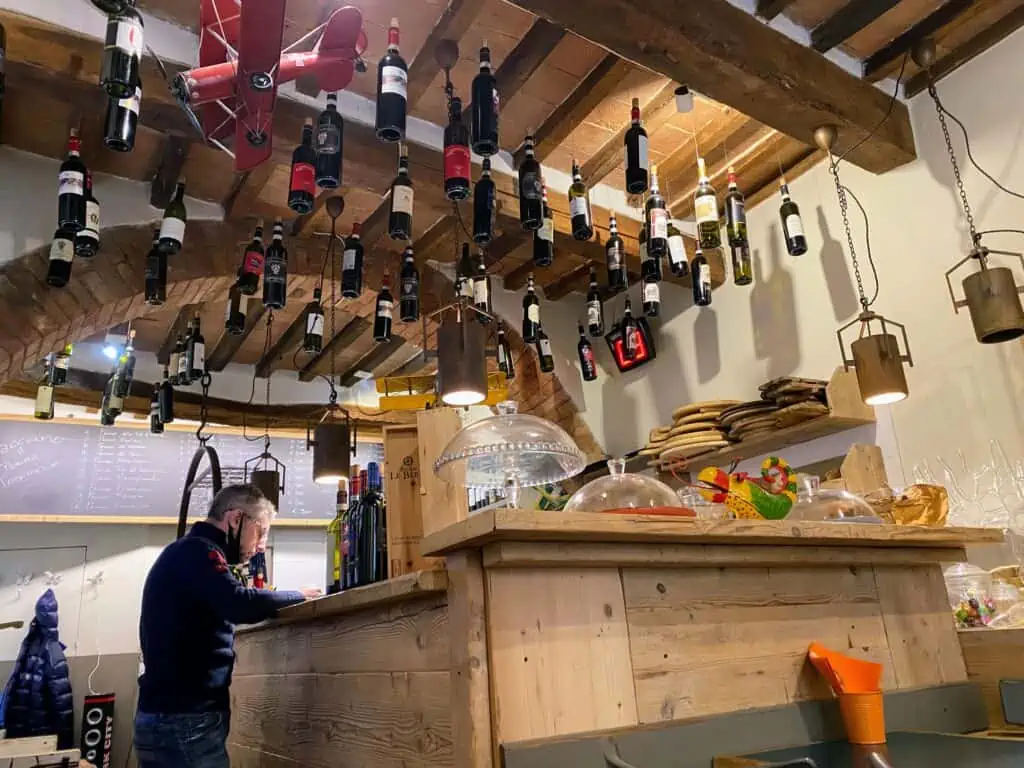 Inside a small restaurant in Italy. Wine bottles hang by strings from the ceiling. Man looks at orders at the counter. Chalkboard menu in back.