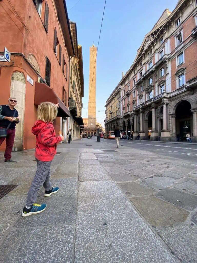 Boy pointing to tall tower in Italian town. It's at the end of the street and there are stone buildings on either side.