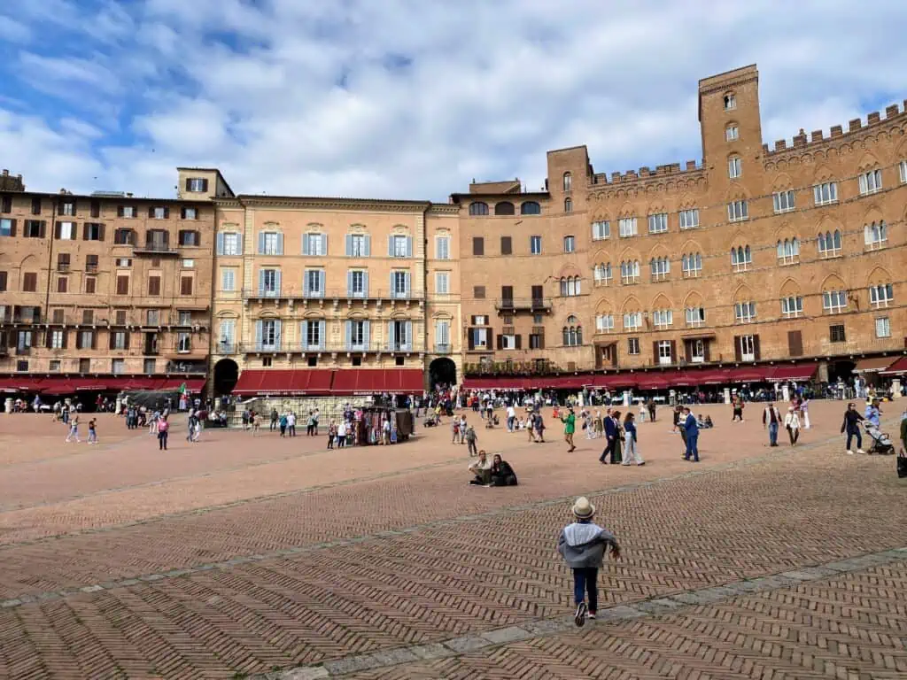 Boys runs across the Piazza del Campo in Siena, Tuscany. There are people sitting in the square and walking in it. You can see buildings and people dining outdoors in the background.