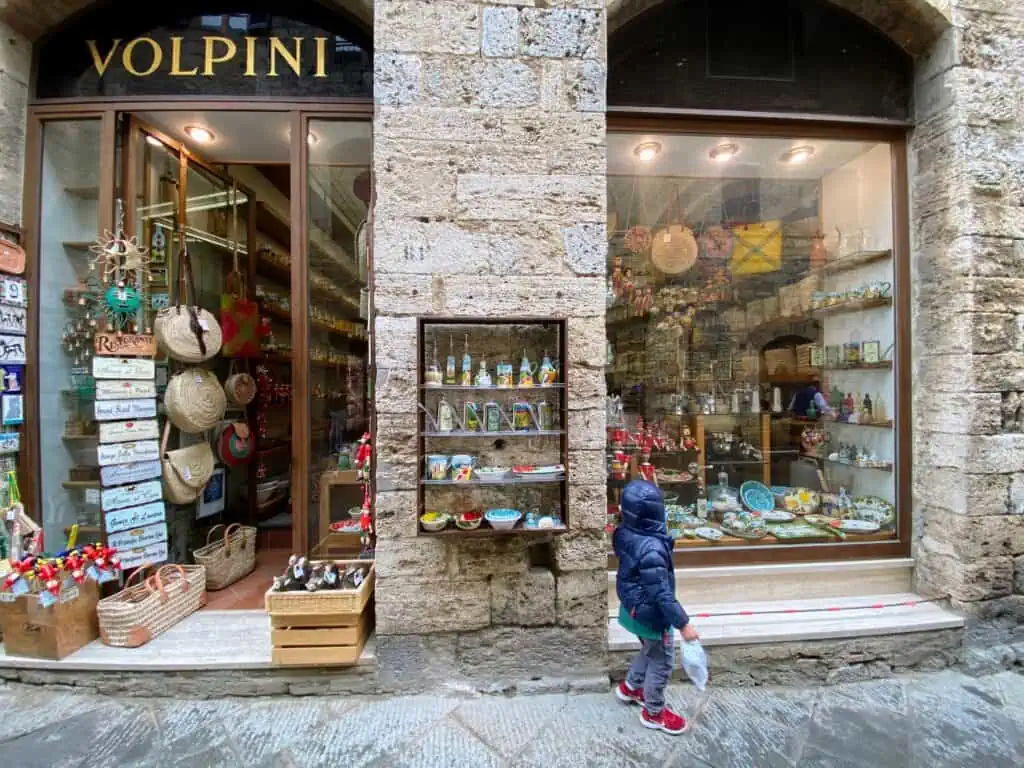 Boy wearing down coat is holding a bag and walking past a shop window in an Italian town (San Gimignano).