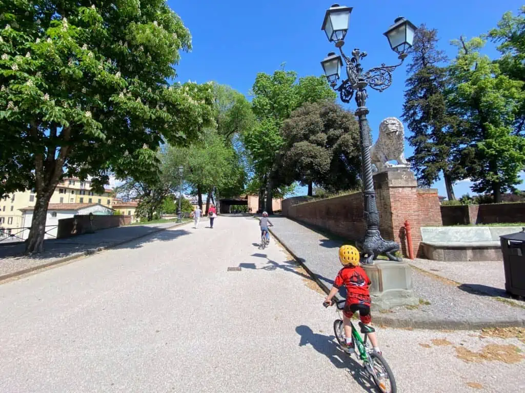 Boys riding bikes on wide path. There is a lamp and stone lion on right and trees on either side of the path. There are two people walking on the path.