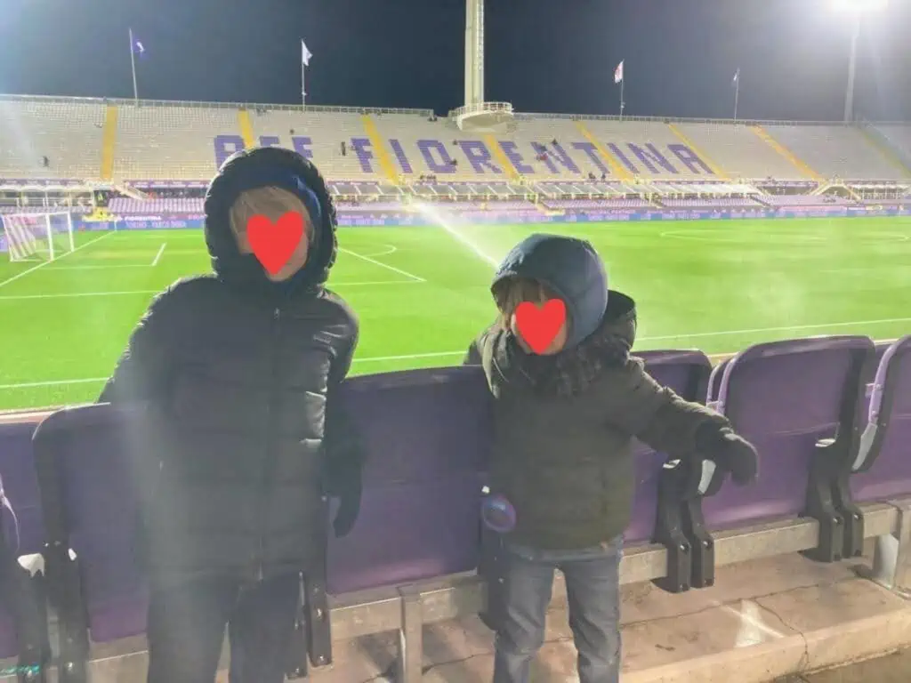 Two boys wearing winter coats and standing by seats at an Italian soccer field. The field is empty.