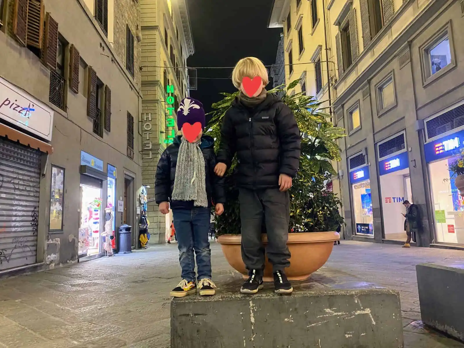 Boys standing on stone block in middle of road in Florence, Italy. You can see closed shops on either side. It's night. They're wearing winter clothing.