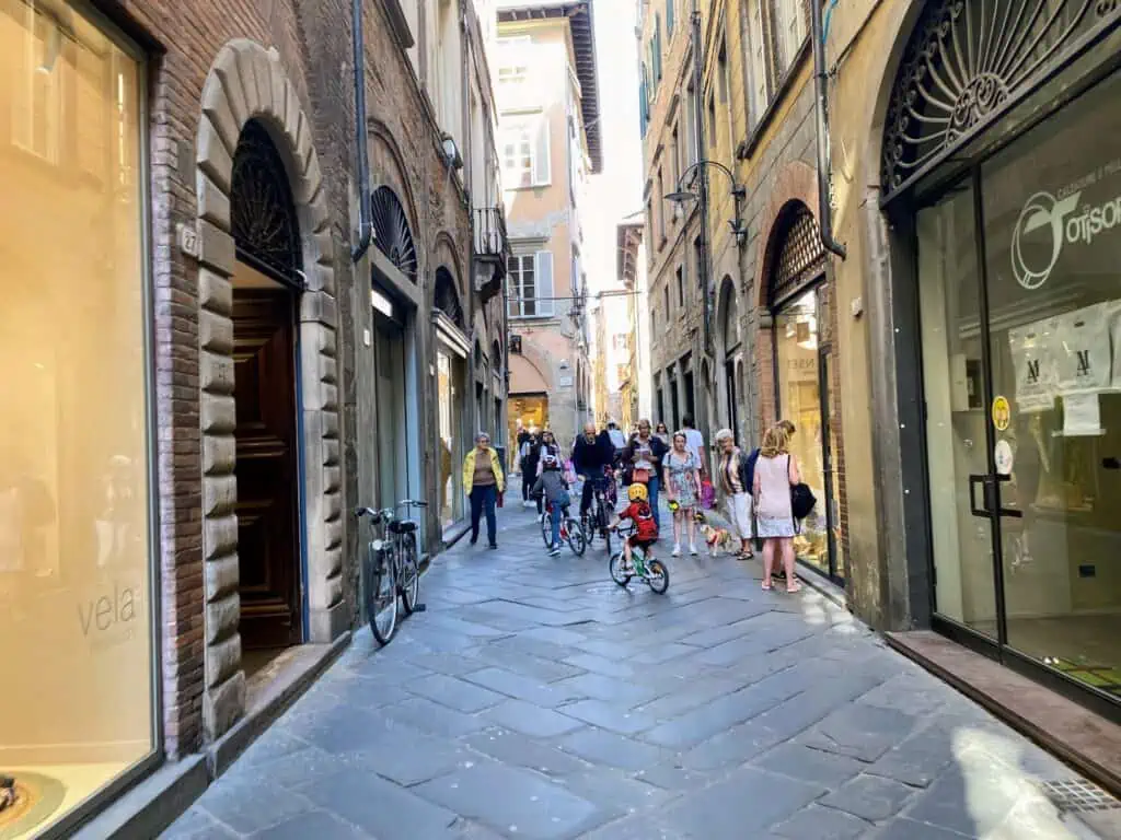 Boys cycling on small street in Lucca, Italy. There are shops on either side and people walking and riding on the same street.