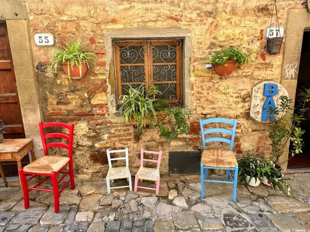 Colorful wooden and straw chairs decorate the front entrance of the bar in Montefioralle, Tuscany. The wall is stone and plants also decorate the wall.