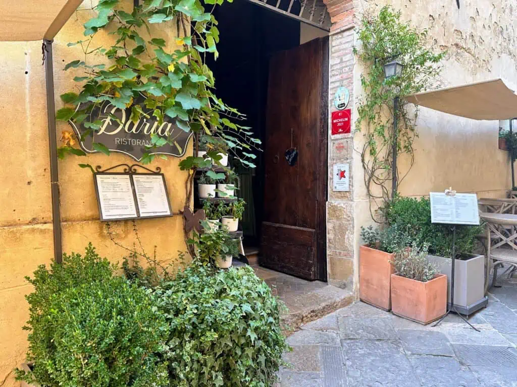 View of front entrance of Daria Ristorante in Monticchiello, Italy. Vines and plants decorate the entrance. You can see the red Michelin sign on the right as well as tables and chairs under a tent.