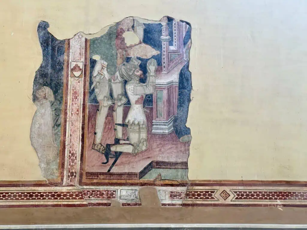 Part of a fresco inside a church in Tuscany. You can see a knight kneeling and praying. Most of the fresco is destroyed and the rest of the wall is cream colored.
