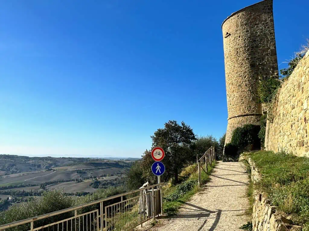 Gravel path leads toward a tall stone tower in a medieval village in Tuscany. On left you can see rolling hills of countryside fields and cypress trees.
