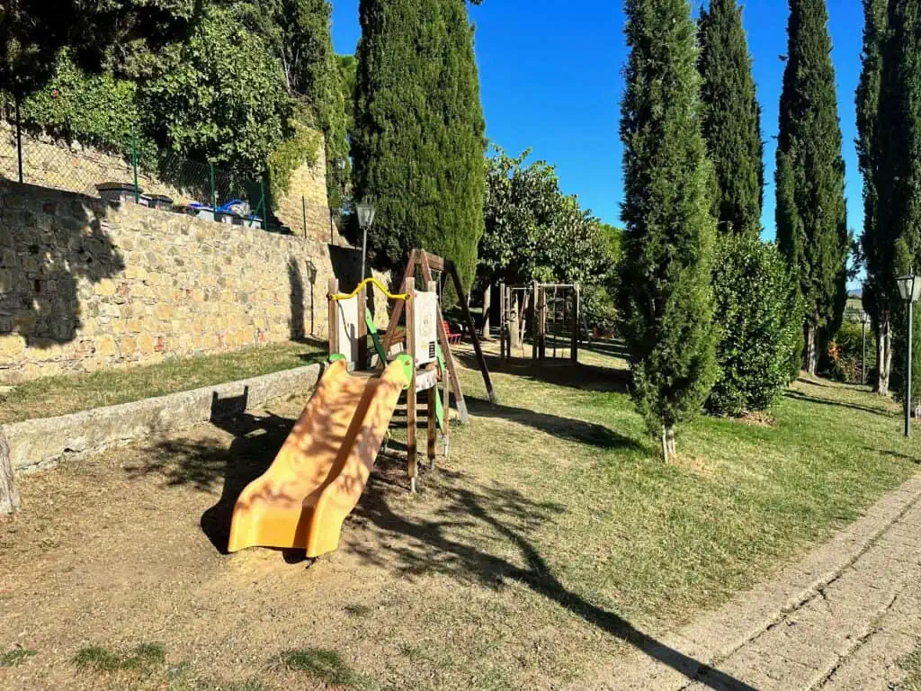 Children's playground with yellow slide and toys. Grassy area surrounded by tall cypress trees. Sunny day with blue sky.