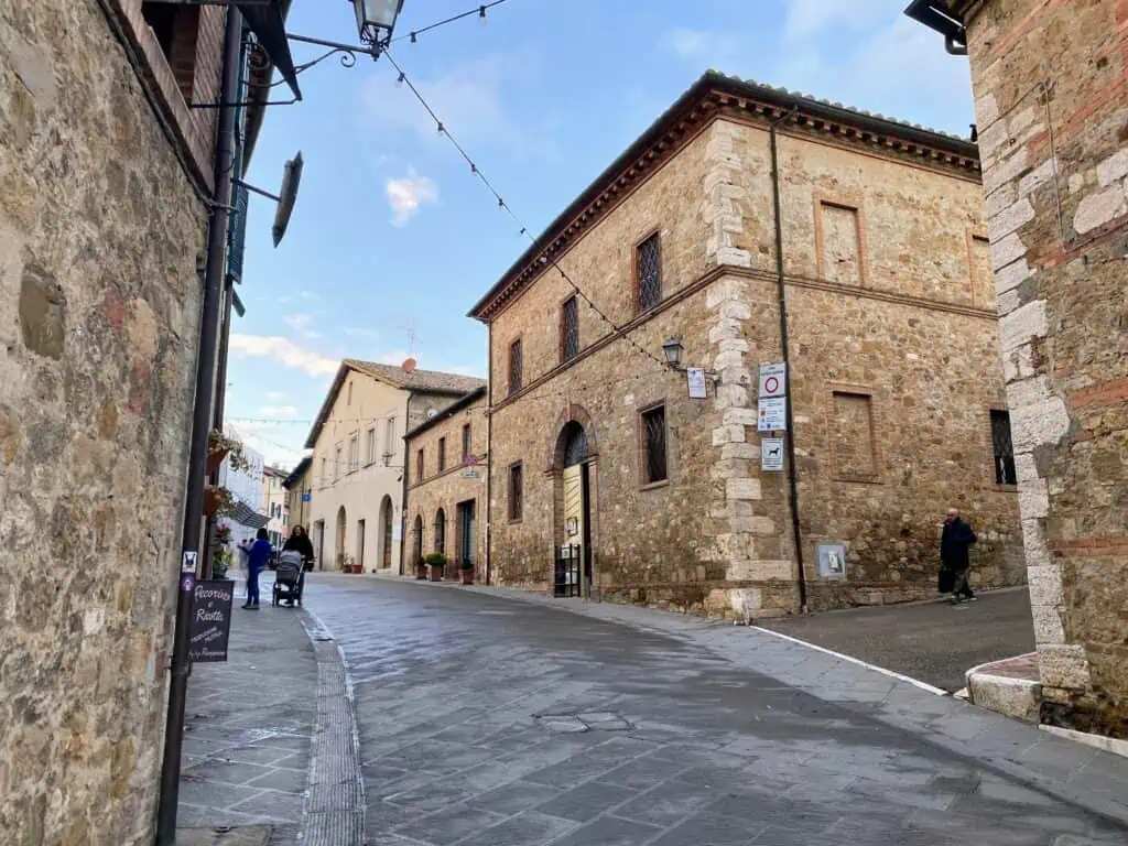 Empty street in a small village in the Tuscan countryside. You can see stone buildings on either side of the street and a few people out walking. They're dressed in winter clothing.