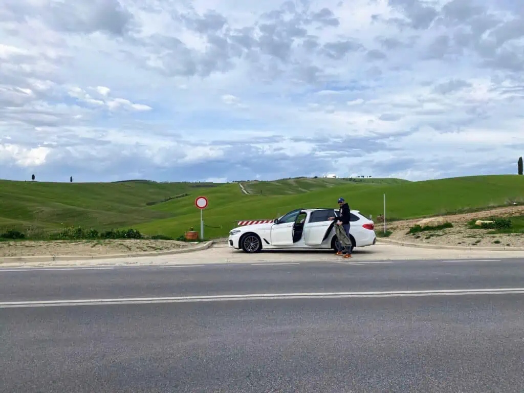 Man standing by rear door of white station. It's parked on the side of the road. In the background are gently rolling green hills and a cloudy sky.