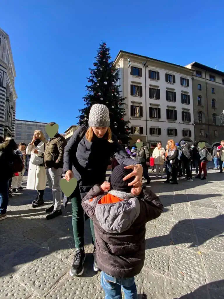 Mother adjusting hat on child in an Italian piazza with a tall Christmas tree in the background.