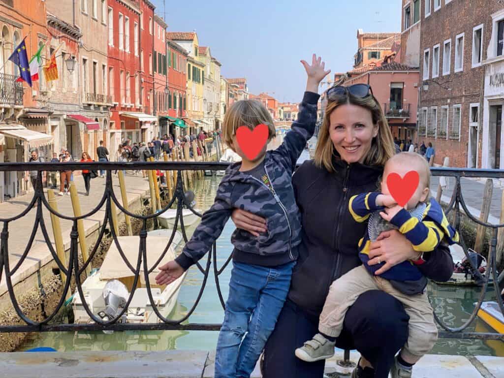 Mom holds sons on bridge. Metal railing behind them as well as a canal with boats and colorful buildings on each side of canal.