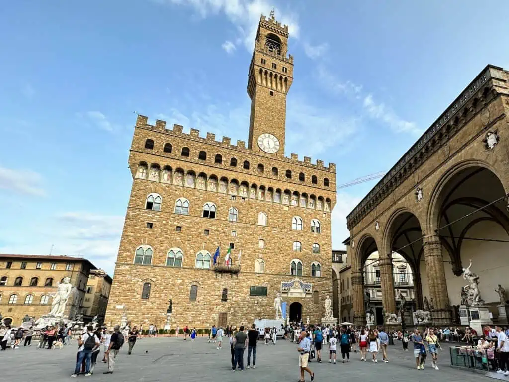 Front view of large stone building with clock tower rising from the center. People walk in the piazza in front and there is a loggia with art on the right.